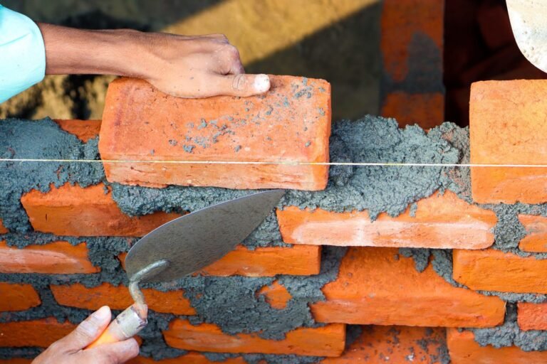 Bricklayer pressing a red brick into a fresh bed of mortar along a string line, trowel in the other hand