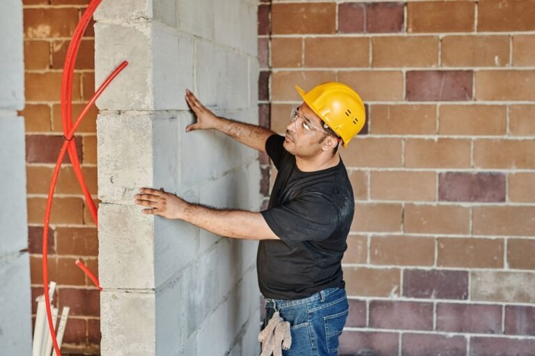 Builder in a yellow hard hat and safety glasses pressing both hands against a grey block wall next to brickwork