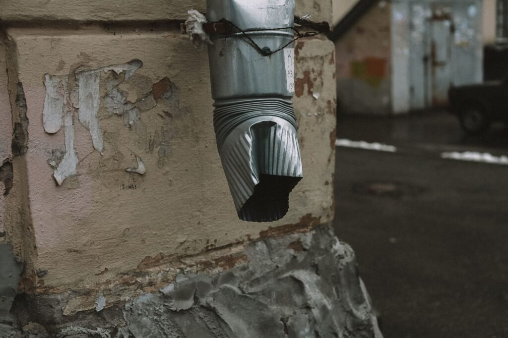 Metal downpipe ending just above the base of a wall with peeling paint and patched, crumbling render