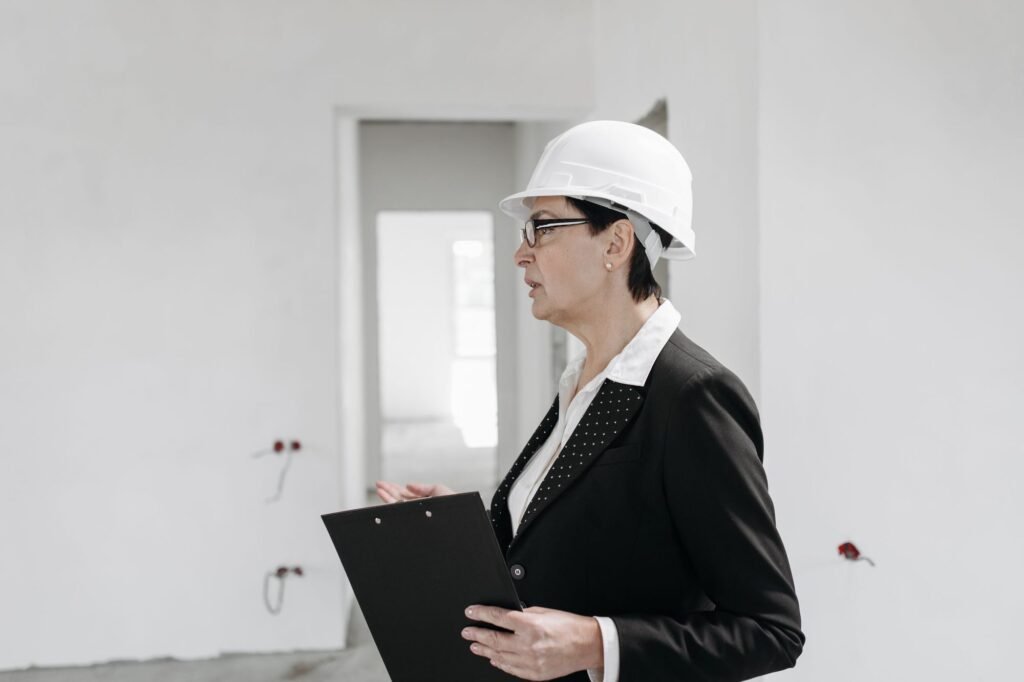 Woman in a white hard hat holding a clipboard while inspecting an unfinished room with bare walls