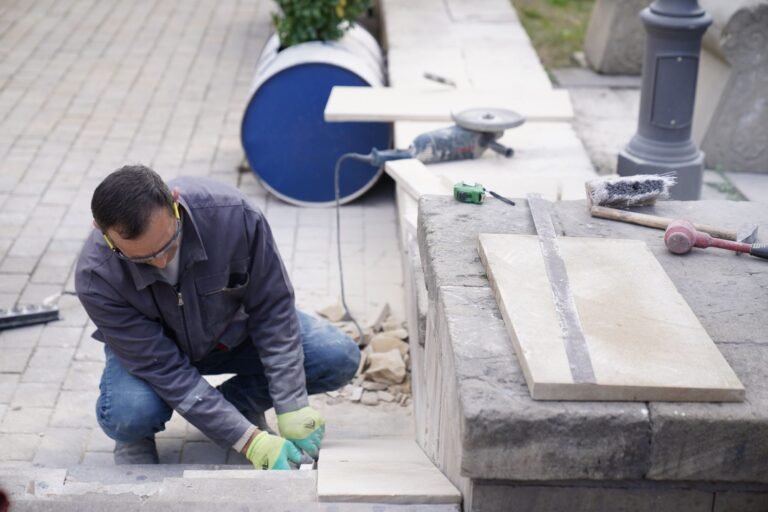 Paver kneeling to fit a sandstone slab on a garden step, with an angle grinder and tools laid on the wall