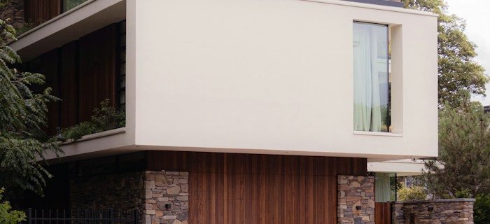 Modern house with a white overhanging upper floor above vertical timber cladding and stone walls, with trees on either side