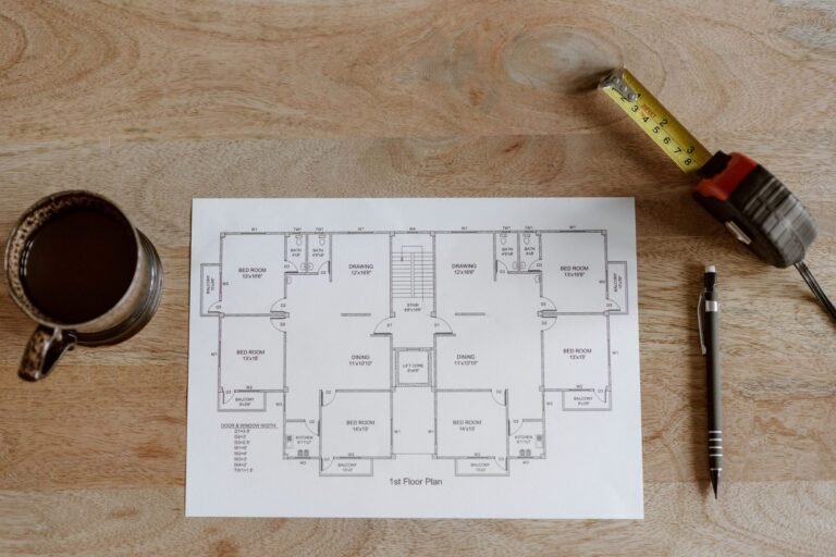 Printed first-floor plan on a wooden table beside a mug of coffee, a pencil and a tape measure