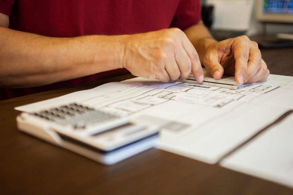 Hands using a scale ruler on a printed floor plan, with a calculator in the foreground