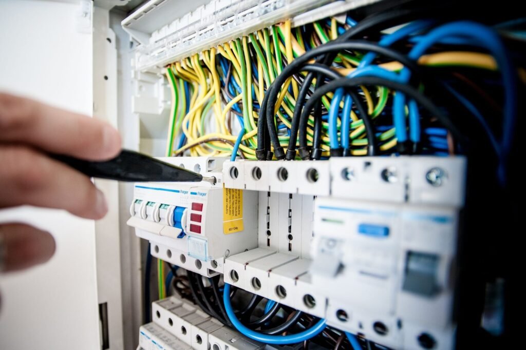 Hand holding a screwdriver at an open consumer unit with a row of breakers and colour-coded cables