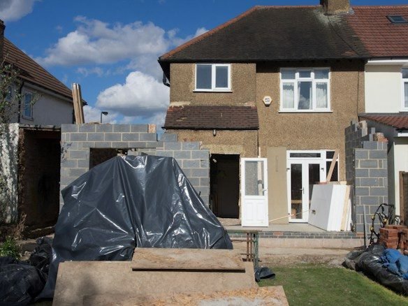 Back of a pebbledashed semi-detached house with part-built blockwork extension walls and a heap under black plastic sheeting