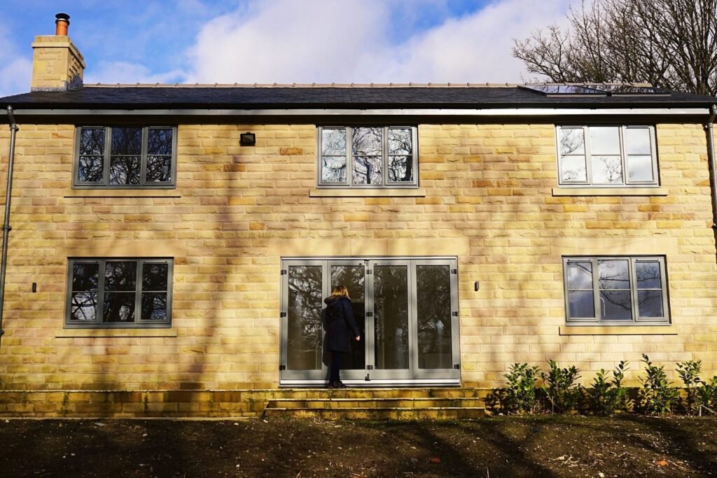 Two-storey house with honey-coloured stone walls, grey-framed windows and bare soil in front, a person at the glazed doors