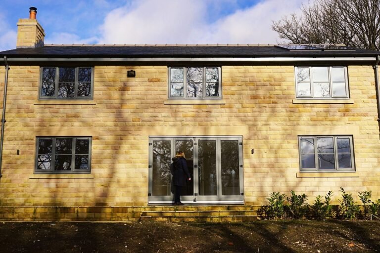 Two-storey house with honey-coloured stone walls, grey-framed windows and bare soil in front, a person at the glazed doors