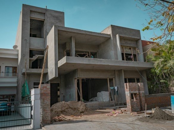Unfinished concrete frame house with open balconies, white sacks stacked inside, heaps of earth and brick piers in front